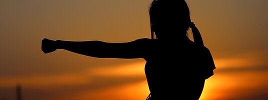 Die Silhoutte einer Frau in einer Tai Chi Pose vor einem Sonnenuntergang.